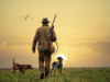 Cazador con escopeta y dos perros de caza caminando por un campo al atardecer con aves volando al fondo